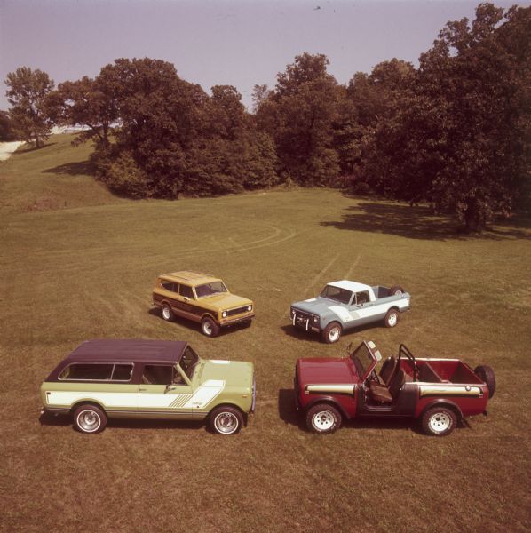 Elevated view of four Scouts parked in a field. On the left is a green Scout with black top, white detailing on side, and the name: "rallye" on front panel in front of passenger door. Top left is an orange Scout with striping on the sides and a roof rack. Top right is a blue Scout with a spare tire in the truck bed, white cab roof, and white detailing on side with the name: "rallye" on front panel in front of driver's side door. Bottom right is a red Scout with black and gold detailing, no roof, windshield, roll bar and spare tire mounted on back.