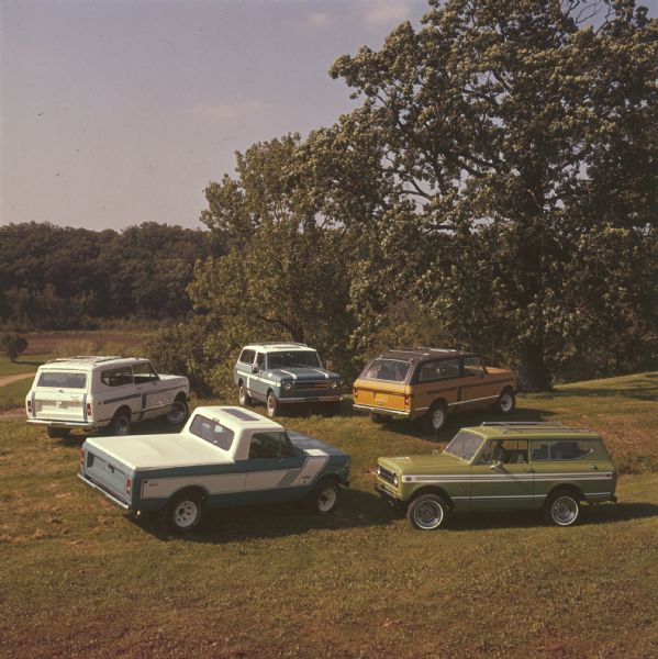Elevated view of five Scouts parked in a field near trees. Bottom left is a blue Scout with white cab, white truck bed cover, and white detailing on side, and the name: "rallye" on front panel in front of passenger door. Top left is a white Scout with white cab roof, roof rack, dark striping on sides. Center top is a blue Scout with white cab roof, roof rack, white striping on sides. Top right is an orange Scout with brown cab roof, roof rack, brown striping on sides. Bottom right is a green Scout with roof rack, white striping on sides.