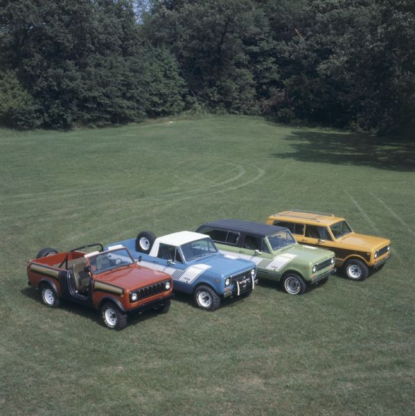 Elevated view of four Scouts parked in an angled row. On the left is a red Scout with black and gold detailing, no roof, windshield, roll bar and spare tire mounted on back. Second from left is a blue Scout with a spare tire in the truck bed, winch on front bumper, white cab roof, and white detailing on side with the name: "rallye" on front panel in front of the passenger side door. Third from left is a green Scout with black top, white detailing on side, and the name: "rallye" on front panel in front of passenger door. On the right is an orange Scout with striping on the sides and a roof rack.