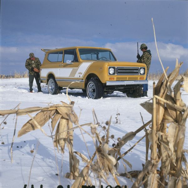 View through corn of Scout parked in the snow. Two men in camouflage coats and hats are standing next to the Scout, and one man is holding a firearm. The Scout is yellow with white detailing on the sides, and the name: "rallye" on front panel in front of driver's side door. The back hatch is open.