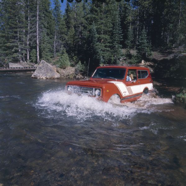 Three-quarter view from front of driver's side of red Scout II with roof rack and white detailing, with the name "rallye" on front panel in front of driver's side door. A man is driving the Scout II across a river in deep water. Trees and boulders are along the shore, and on the far left is a wooden bridge.