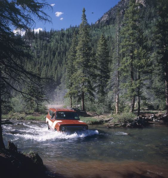 View from shoreline of Scout II being driven across a river. Trees,  hills and a mountain are in the background. The red Scout II has a roof rack and white detailing, with the name "rallye" on front panel in front of passenger side door.