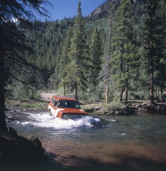 View from shoreline of Scout II being driven across a river. Trees, hills and a mountain are in the background. The red Scout II has a roof rack and white detailing, with the name "rallye" on front panel in front of passenger side door.