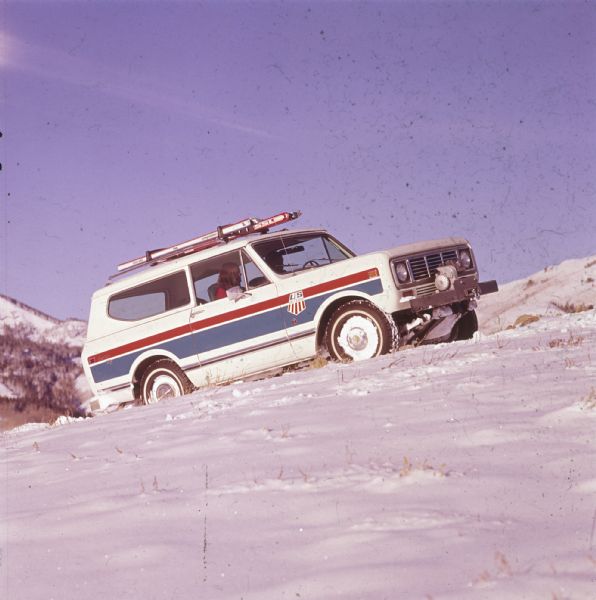 Passenger side view of a white Scout II XLC with red and blue detailing on the side. The Scout is being driven up a snowy hill in the mountains. A woman is sitting in the passenger seat, and the US Ski Team emblem is on the side panel in front of the door. Skis are attached to the roof rack, and there is a winch with a hook on the front bumper. Trees and mountains are in the background.