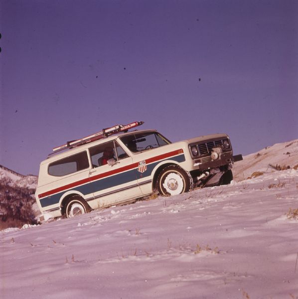 Passenger side view of a white Scout II XLC with red and blue detailing on the side. The Scout is being driven up a snowy hill in the mountains. A woman is sitting in the passenger seat, and the US Ski Team emblem is on the side panel in front of the door. Skis are attached to the roof rack, and there is a winch with a hook on the front bumper. Trees and mountains are in the background.