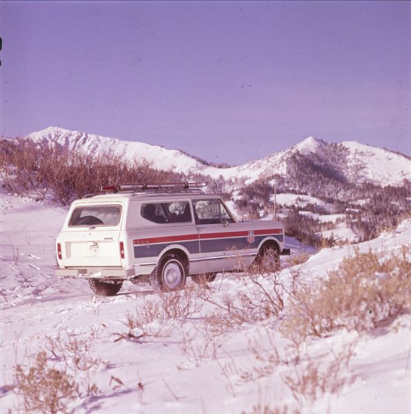 Three-quarter view from rear of passenger side of white Scout II XLC. The Scout is being driven on a snowy path in the mountains. A man appears to be driving, and a woman sits in the passenger seat. The US Ski Team emblem is on the side panel in front of the door. Skis are attached to the roof rack. Mountains are in the background.