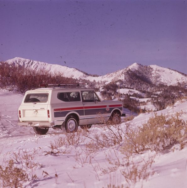 Three-quarter view from rear of passenger side of white Scout II XLC. The Scout is being driven on a snowy path in the mountains. A man appears to be driving, and a woman sits in the passenger seat. The US Ski Team emblem is on the side panel in front of the door. Skis are attached to the roof rack. Mountains are in the background.