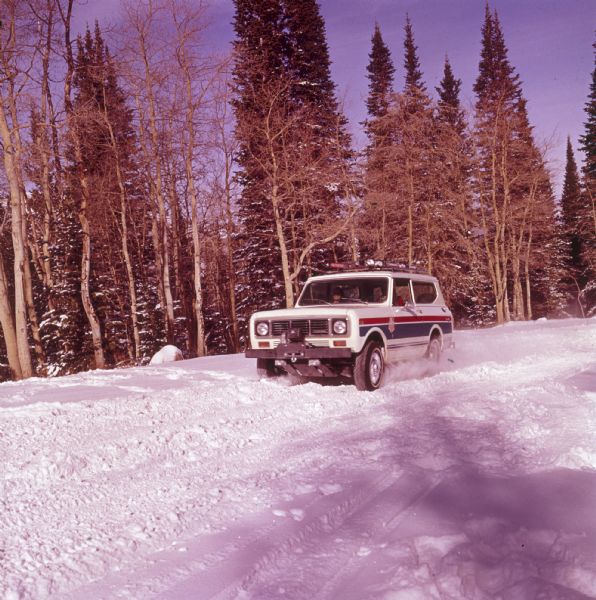 Three-quarter view from front of driver's side of white Scout II XLC. The Scout is being driven on a snowy road with trees in the background. A man appears to be driving, and a woman sits in the passenger seat. The US Ski Team emblem is on the side panel in front of the driver's door. Skis are attached to the roof rack, and a there is a winch with a hook on the front bumper.