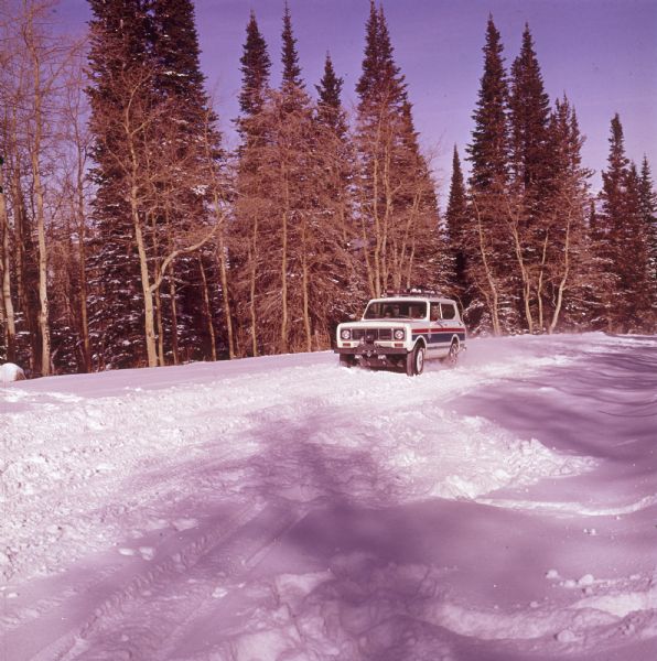 Three-quarter view from front of driver's side of white Scout II XLC. The Scout is being driven on a snowy road with trees in the background. A man appears to be driving, and a woman sits in the passenger seat. The US Ski Team emblem is on the side panel in front of the driver's door. Skis are attached to the roof rack, and a there is a winch with a hook on the front bumper.
