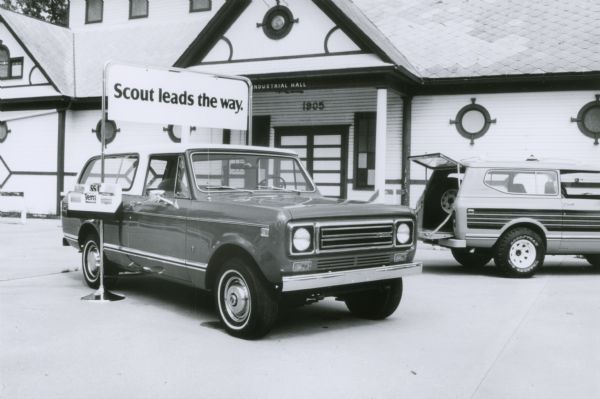Scout parked outdoors underneath a sign that reads: "Scout leads the way." There is a box of literature attached to the pole that advertises Terra and Traveler Scouts. Another Scout is parked in the background on the right. A large building in the background has a sign above the entrance that reads: "Industrial Hall" and a date of 1905.