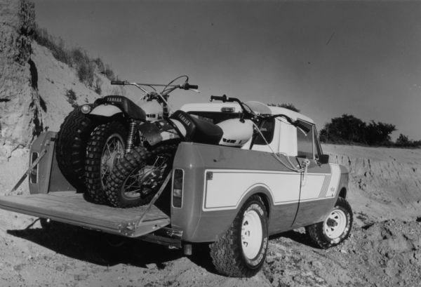 Three-quarter view from rear of passenger side of Scout pickup, which is parked in a sandy area with dunes, loaded with two Yamaha motorcycles. The Scout has white detailing, with the name "rallye" on front panel in front of passenger side door. The tailgate of the pickup is open.