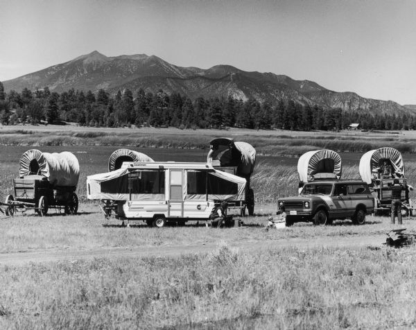 Scout and a Coachmen pop up camping trailer parked in a field in a valley. A row of covered wagons are behind the Scout and the camping trailer. In the background are trees and a mountain range. A man on the right is taking a photograph of a man holding a rifle posing in one of the covered wagons. A woman sits on the ground with an infant, and a man squats near a cooler.