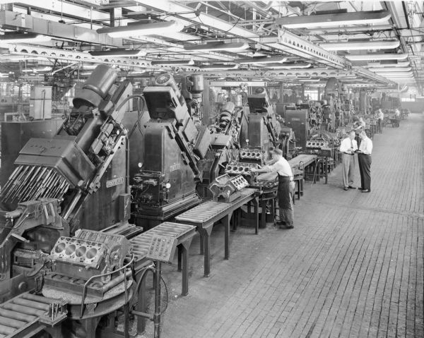 Men working at machinery on a large factory floor. Engine blocks are on conveying belts. The name on the machinery is "Footburt." Two men, both wearing eyeglasses and neckties, stand in the aisle talking.