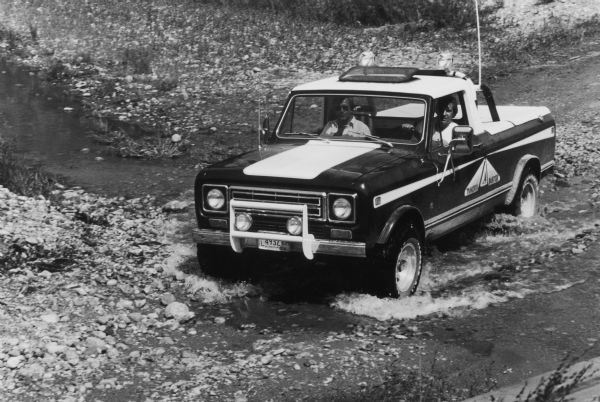Two men in a Scout are driving off-road through a stream.