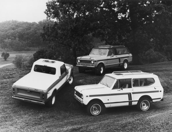 Three Scouts parked outdoors in a field near trees. The Scout II on the left has a cover over the truck bed, and a sunroof on the top of the cab. White detailing on side with the name "rallye" on front panel in front of passenger side door. The Scout in the back has a roof rack and a sunroof, and there is dark detailing on sides and top of hood. The Scout on the bottom right has dark detailing on sides and top of hood, a roof rack, and a sunroof.