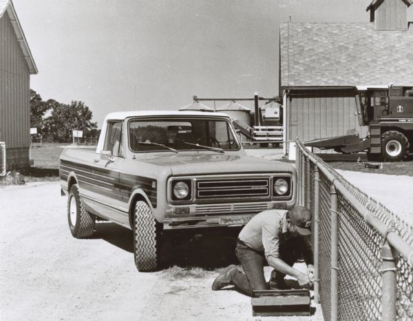 An unidentified man is kneeling to repair a chain link fence. Parked behind him is an International Scout II. In the background is an International 1460 combine with a corn header, suggesting that this is a farm.