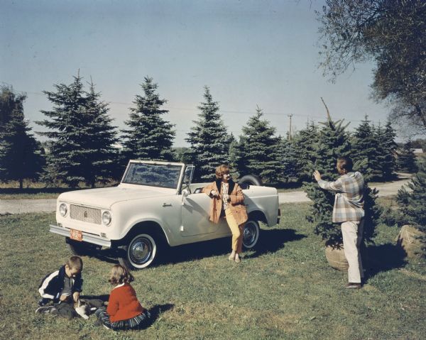 A man, woman, and two children posing with a white 1963 Scout 80. The spare tire is mounted on the back. There is no top on the Scout. The man is standing and holding an evergreen tree, and the two children are sitting on the ground with a cat. The woman is leaning against the driver's side door of the Scout.