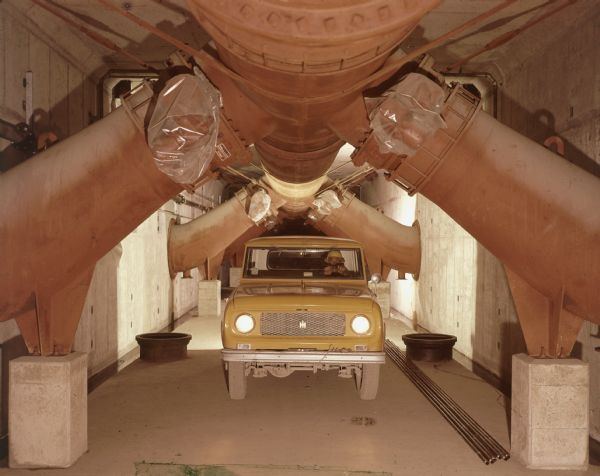 View from front of a man driving a yellow Scout inside a cavernous industrial tunnel underneath large pipes.