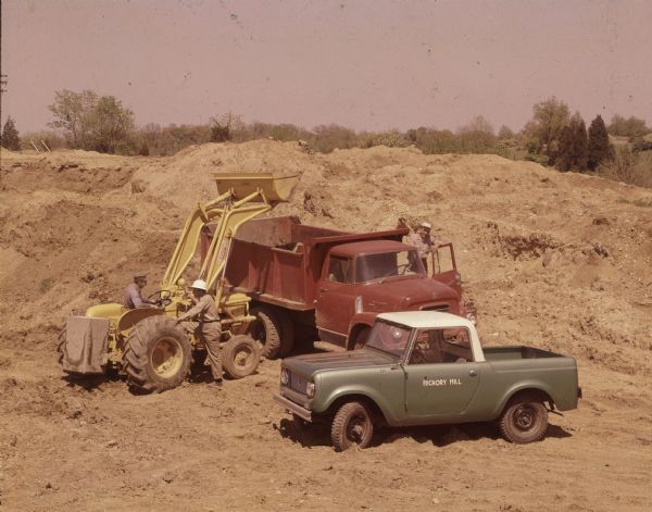 Slightly elevated view of three men posing with a Scout pickup, International dump truck, and International/Wagner front end loader. On the driver's side door of the Scout is painted: "Hickory Hill." Around the men are piles of dirt, and in the far background are trees.