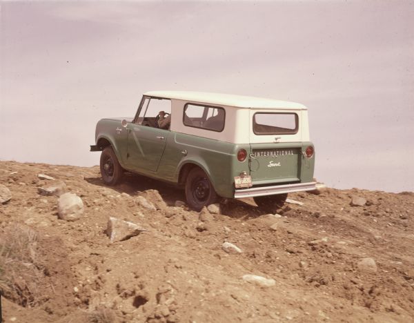 Three-quarter view from rear of driver's side of a green Scout pickup with white topper. A man and a passenger are traveling up a rocky incline.