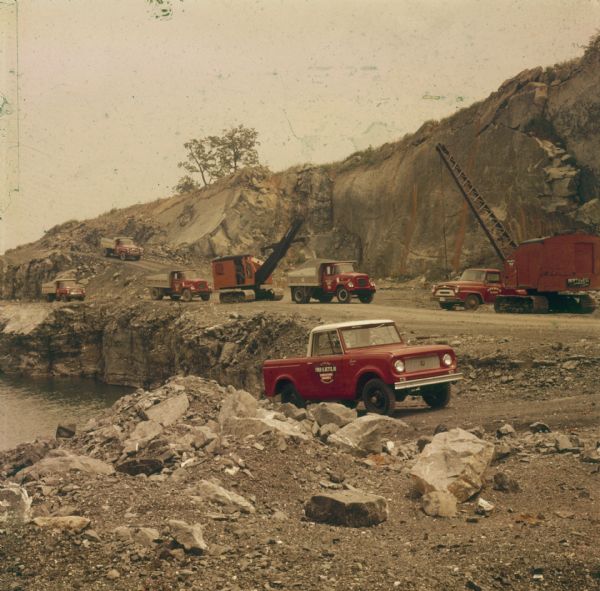 Man driving a red Scout pickup with white cab top. Side of passenger side door has a sign that reads: "Fred K. Betts, III Limestone Quarry." Dump trucks and cranes and a steep incline are in the background, and water is in the quarry on the left.