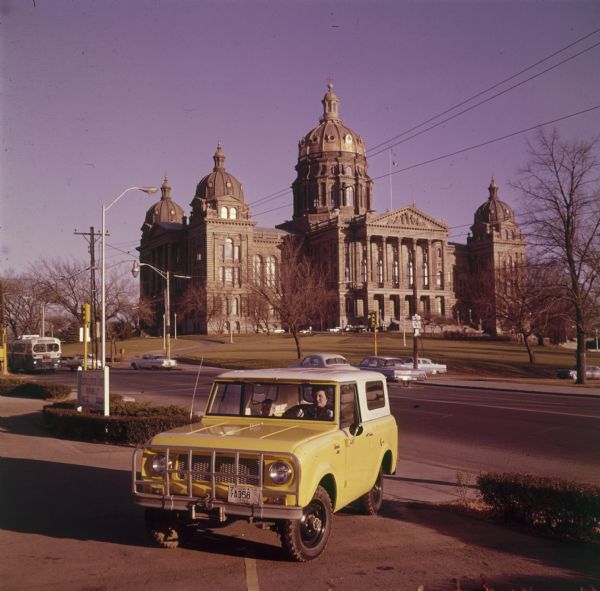 View from front of woman driving a yellow Scout with white topper. There is a young boy in the passenger seat. A grill and tow hook are attached to the front bumper. Across the street in the background is the Iowa State Capitol building.