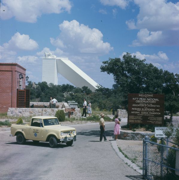 Man driving yellow Scout on road near the visitor center for the Kitt Peak National Observatory. Men and women are standing near the entrance, and the observatory is in the background.