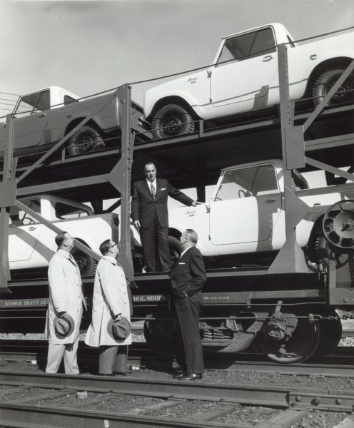 Three men stand on railroad tracks, looking up at another man standing on the bottom level of a railroad car, stacked double-decker style with white Scout pickup trucks.