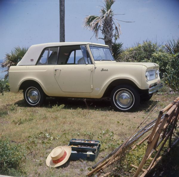 Man sitting in pale yellow Scout parked at bird sanctuary. Hat and fishing gear are resting on the grass in the foreground, ocean is in the background.