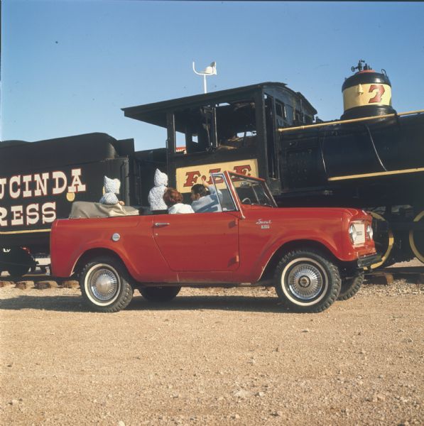 Family in red Scout convertible. The man in the driver's seat is pointing at the locomotive they are parked in front of. A woman is sitting in the passenger seat, and two young children wearing matching jackets with hoods, are standing in the back looking the train.