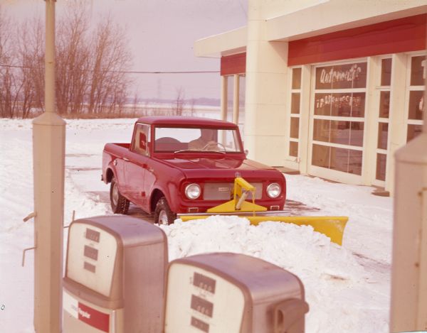 View over gas pumps of a man using an International Scout pickup to plow snow at a Citgo service station. The Scout is painted red, and the snow plow is painted yellow.
