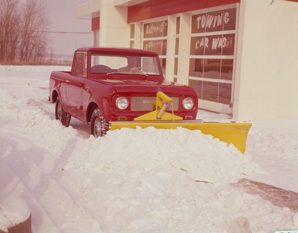 View from front of man driving red International Scout pickup with yellow snow plow on front. He is plowing at a Citgo service station.