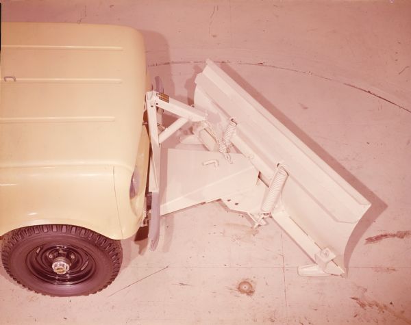 Slightly elevated view of passenger side showing the front wheel and hood of yellow International Scout. A snow plow, painted white and with an "IH" insignia, is attached to the front bumper.