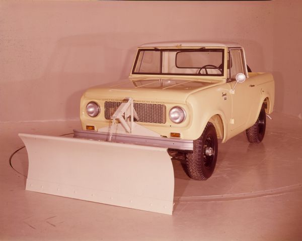 Three-quarter view from front of driver's side of yellow International Scout with a white snow plow attached to the front. The pickup has a white cab top, and a spare tire is mounted on the back of the tailgate.