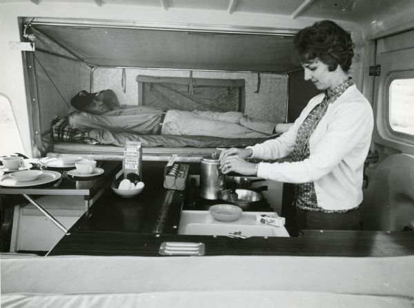 A man reclines on the bed of the extended Scout camper, watching a woman cooking breakfast on the stove. Butter, eggs, milk, coffee and donuts are visible along with dishes. 