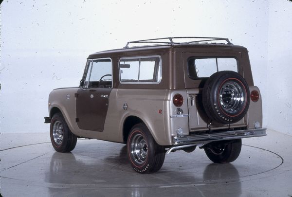 Three-quarter view from rear of driver's side of International Scout Aristocrat. Light brown and dark brown body and topper. There is a roof rack, and a spare tire is mounted on the back door.