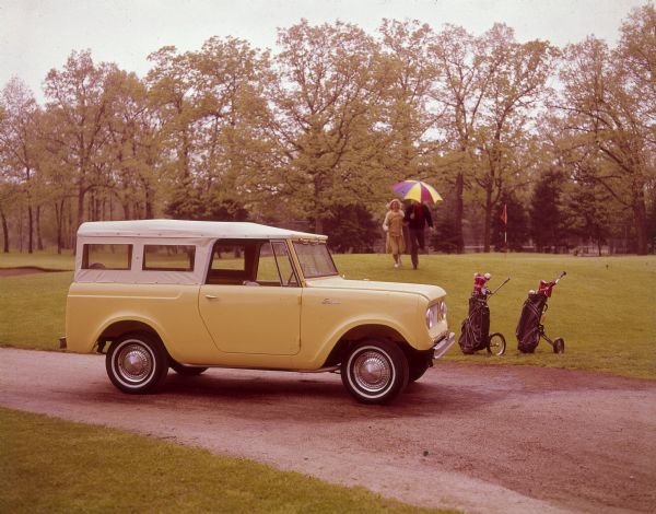Yellow Scout with canvas roof parked near a golf course with two golf bags nearby. A couple is running towards the Scout. The man is holding an open umbrella.