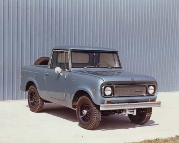 Three-quarter view from front of passenger side of blue Scout 4x4 parked outdoors. The truck bed is open. A spare tire is mounted on the back of the tailgate. A metal sided building is in the background. The driver's side window and rear window of cab have been covered with white material.