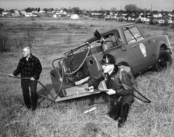 Two men with fire hoses are standing in a field next to a Scout carrying fire equipment. The Scout is parked on a hill.