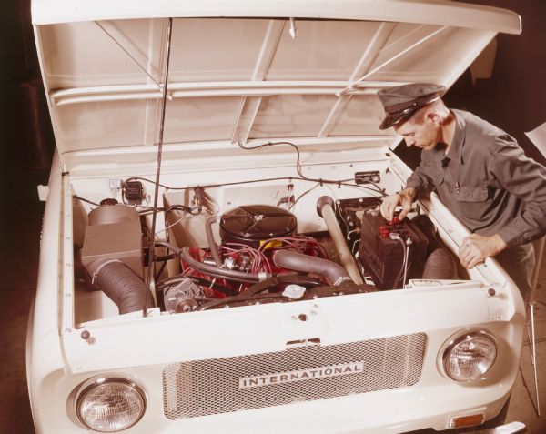 A man wearing a uniform and a hat is shown inspecting the battery under the hood of a white Scout 4x4.
