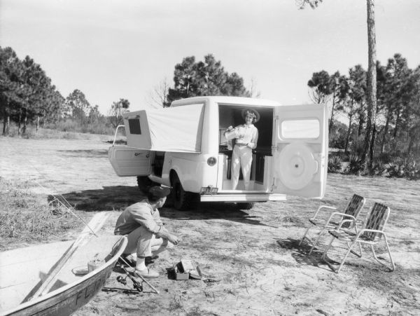 International Scout De Luxe Camper promotional image. A woman is standing at the open back door pouring a cup of coffee. A man is looking up at her while working with fishing equipment, he is crouched on the ground near a boat. The camping scene is in a wooded area. Original caption reads: "Open the back door of a new Scout de luxe Camper by International and you step onto a modern compact home on wheels. The tented 'wings' of the house are foam-padded beds. Inside is a stand-up galley complete with range sink, refrigerator and an adjoining dinette table and benches. A chemical toilet hidden in the wall can be pulled out and screened from view. There are screened windows in the bedroom wings, and screens can be obtained for the Scout front door windows. Also available is a canopy to form a roofed patio at the Scout Camper's back door. Power for the all-wheel drive Scout Camper is provided by a 93 hp. Comanche engine." 