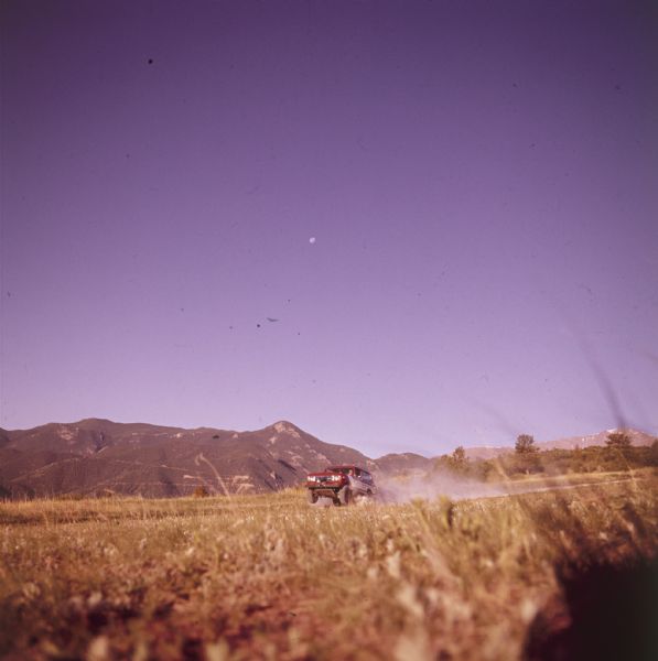 Scout doing a wheelie on a road through a field. Mountains in background, and moon high in the blue sky.