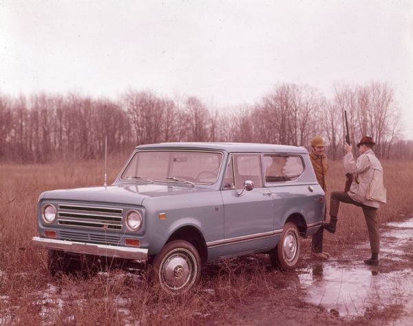 Two men are standing at the back of a blue Scout II with blue topper in a field with snow and ice on the ground. They are wearing hunting jackets and hats, one man is holding a rifle or shotgun.