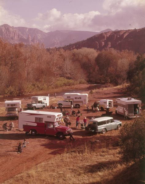Elevated view of group of people with six Scouts, most with campers mounted on the truck beds, parked near a river. Mountains are in the background.