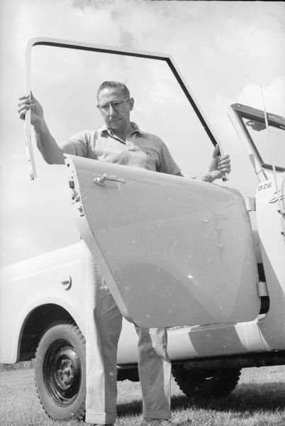 Man holding detached window frame above the open passenger door of a Scout. The number "926" is painted on the side panel just in front of the passenger side door.