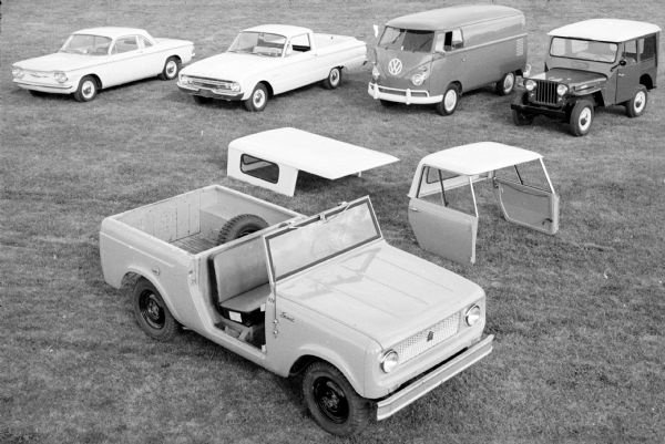 Elevated view of Scout pickup parked outdoors on the grass. The spare tire is mounted in the open truck bed. Behind the Scout sitting on the grass is the truck bed cap, and also the cab roof with two doors. Parked in a row behind the Scout are four vehicles. One is a VW van. Another vehihcle may be a Ford Ranchero.