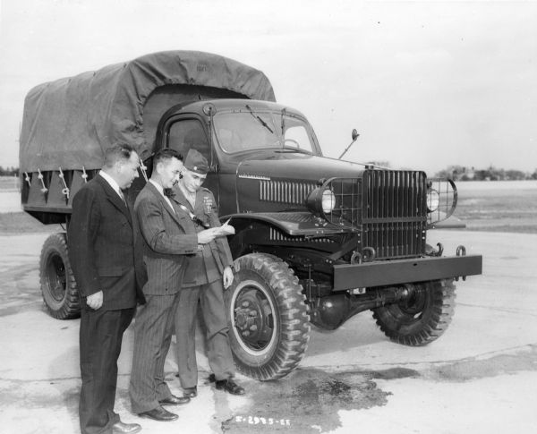 Three men stand outdoors in front of an International military vehicle looking at paperwork. Two of the men are wearing suits, and the third man wears a military uniform.