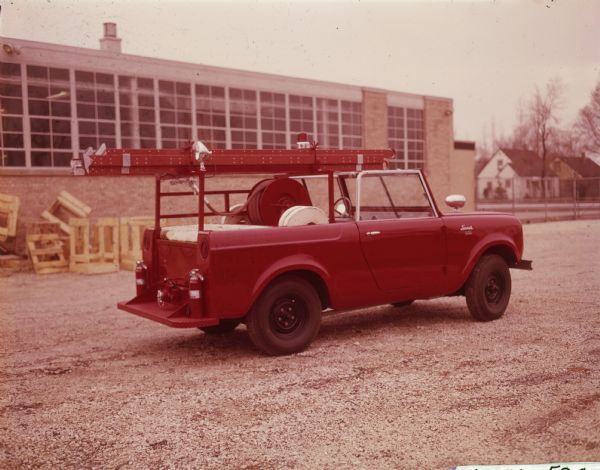 Three-quarter view from rear of passenger side of Mini-Fire truck parked outdoors. A ladder is attached on top of a frame built in the truck bed. Fire hoses are on reels in the truck bed. Two fire extinguishers are sitting on the running board behind the tailgate. Industrial building in the background.