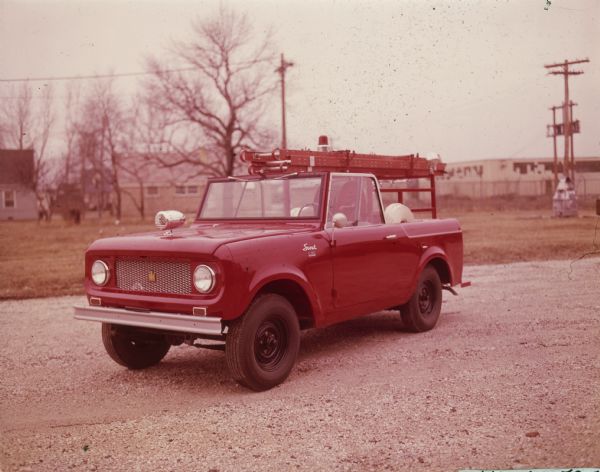 Three-quarter view from front of driver's side of Mini-Fire truck parked outdoors. A ladder is attached on top of a frame built in the truck bed. Fire hoses are on reels in the truck bed. Two fire extinguishers are sitting on the running board behind the tailgate.
