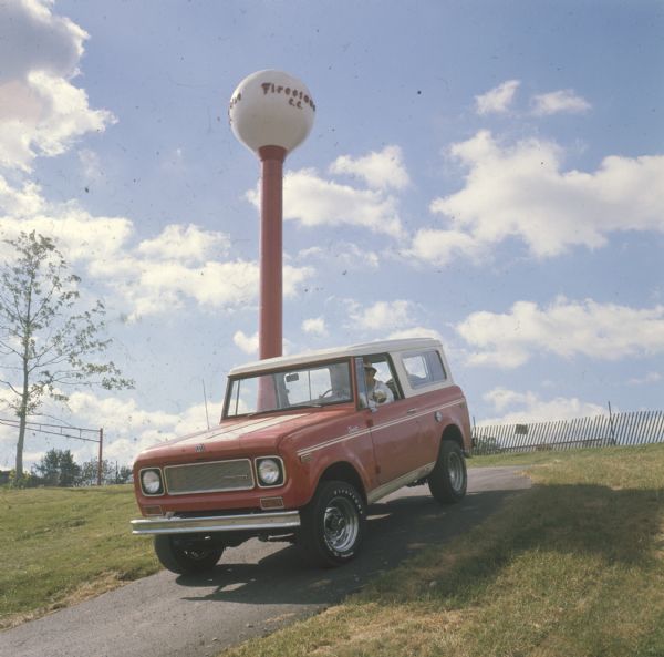 Man driving red Scout with white topper. On the lower side panel near the back is painted, "SR-2." In the background is what may be a water tower, with the sign: "Firestone C.C."
